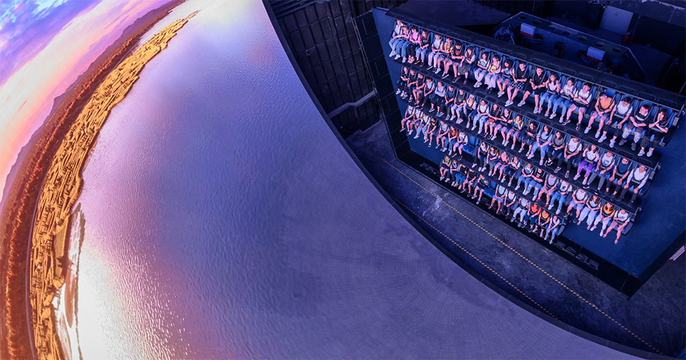 A number of people sit along four rows on a vertical seating system looking towards a large curved screen that gives the impression that they are hovering over the Earth