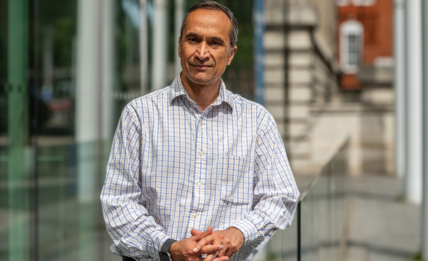 Aman wearing a blue and white checked shirts stands outside and looks at the camera 