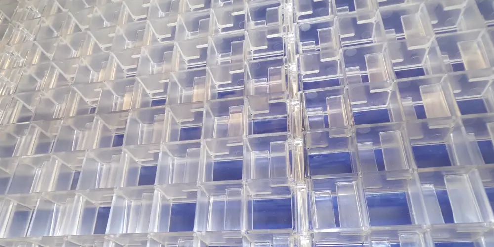 Close-up view of a grid of transparent plastic panels arranged in a repeating pattern. The panels form square compartments with clear edges, creating a geometric, layered effect. A blue surface is visible beneath the grid.