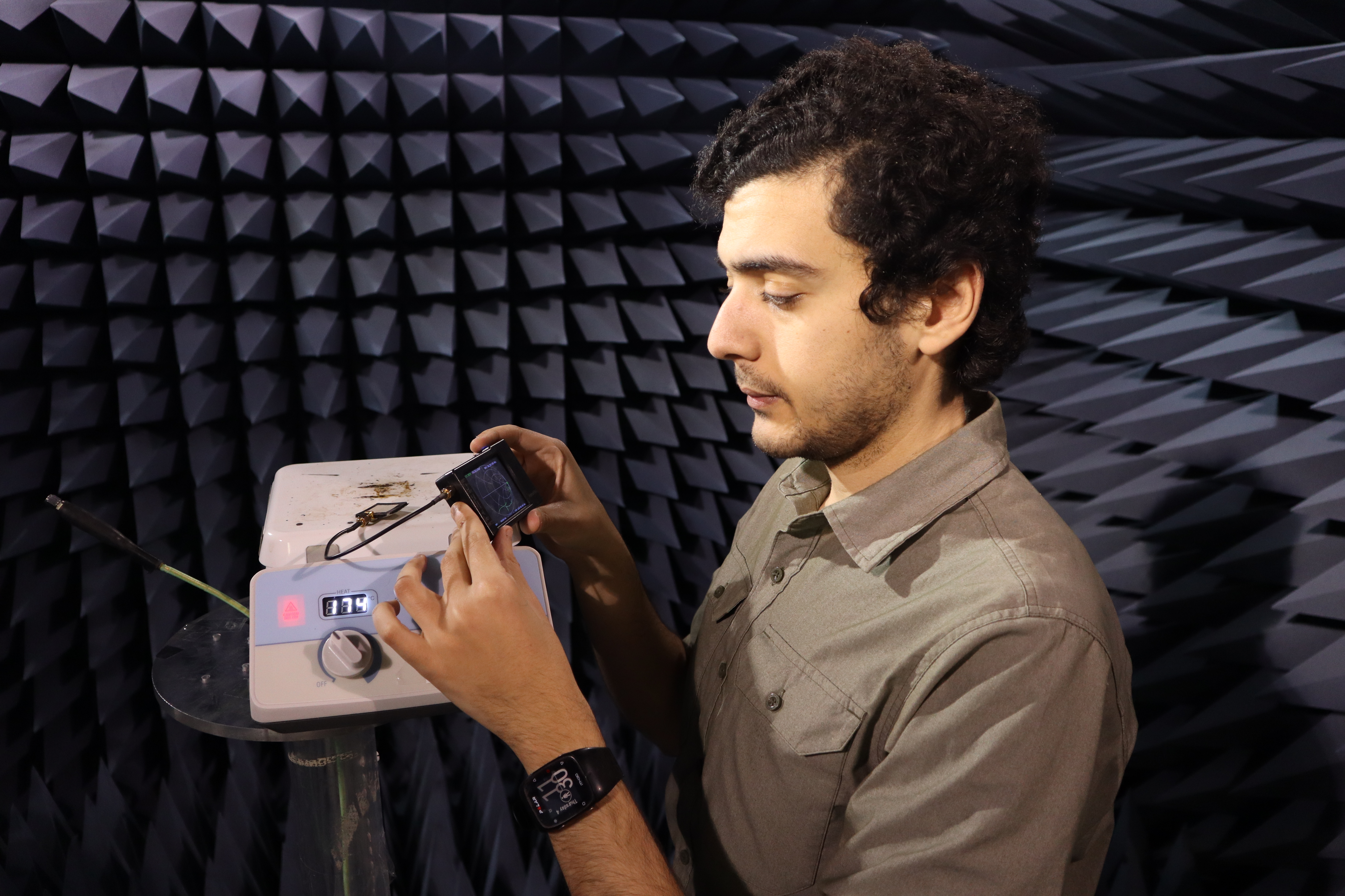A man with brown hair wearing a beige shirt in a soundproof room, looks at an electronic device held in his hands