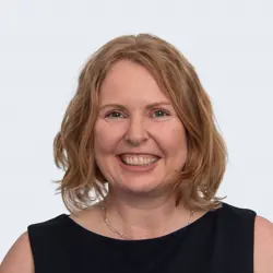 A portrait of a woman with a blonde bob and blue eyes smiling, wearing a black boat neck top.
