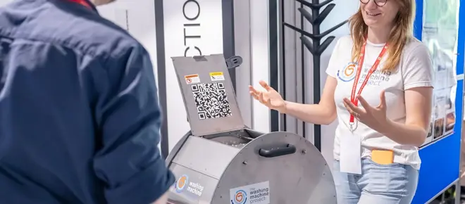 Person explaining a metal drum device during a demonstration, with another attendee listening and display boards in the background.