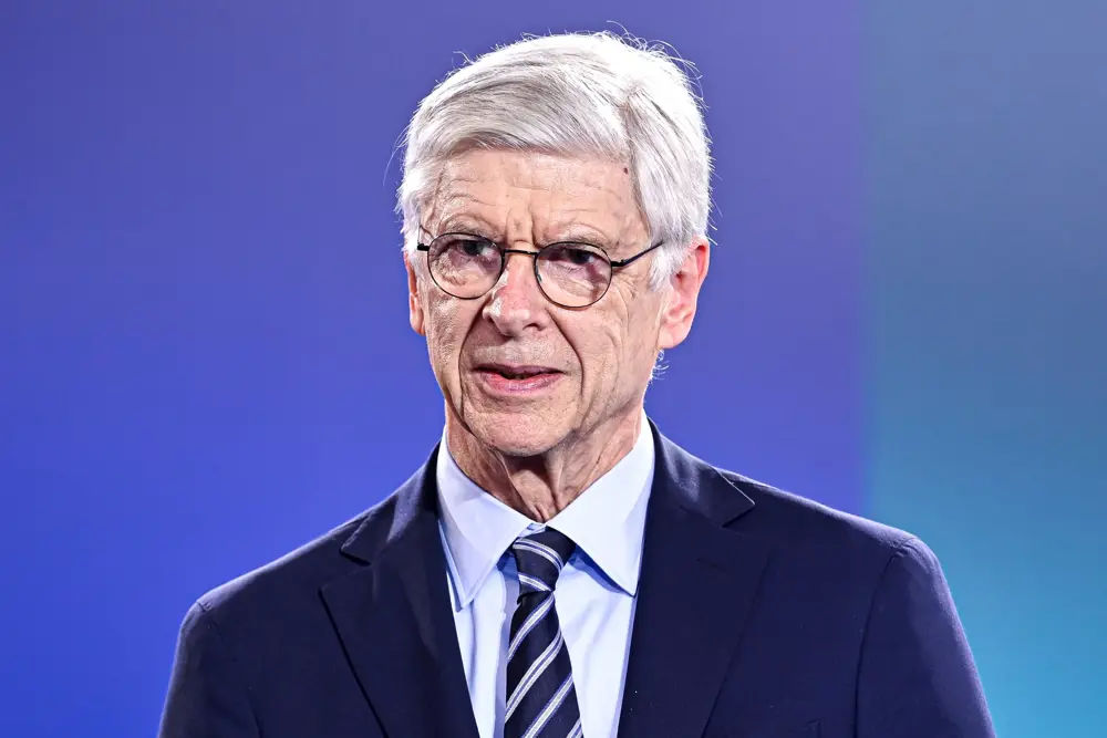 Close up of Arsene Wenger wearing a suit and glasses, speaking at an event, in front of a blue background.
