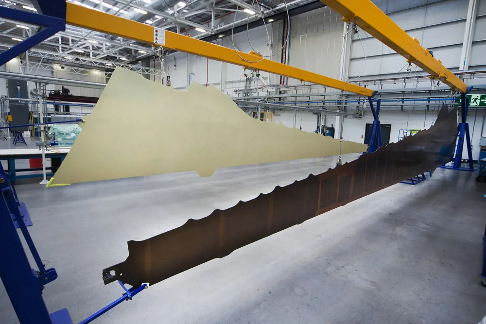 A thin large one piece wing skin hanging from a bar above the ground in a factory.