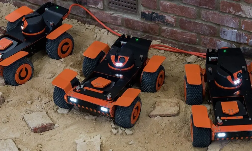 Three models of the orange and black Q-bot Spraybot robots, on a sandy floor next to a brick wall.