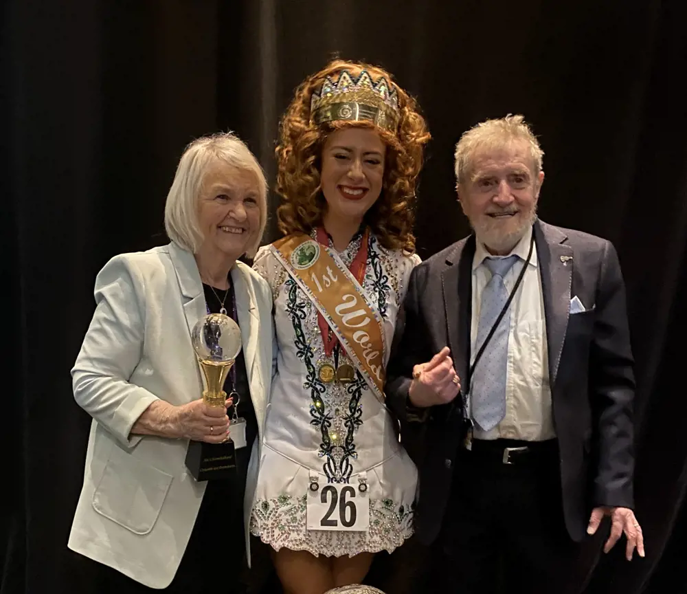 A young woman and two older people smiling for a photograph, the woman is wearing a first place sash and the lady to her left is holding a metal and glass trophy.