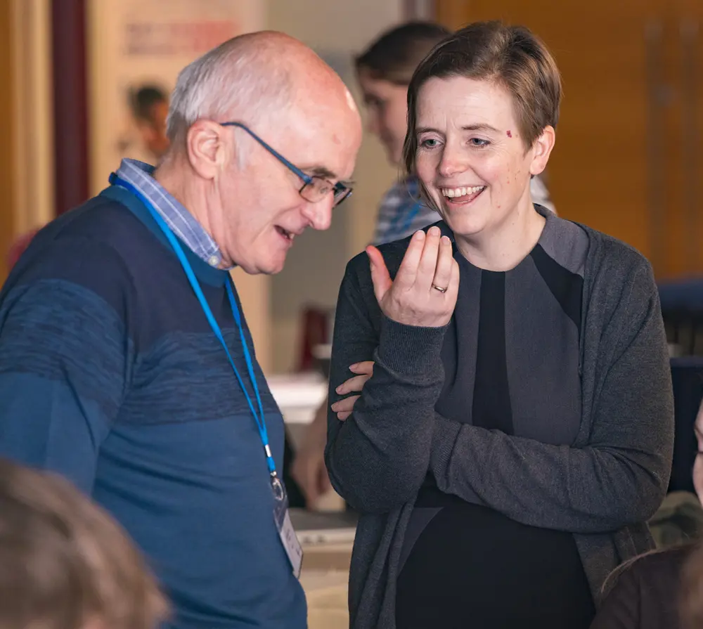 Two people in conversation at an indoor event, one gesturing while the other looks down, both wearing name badges.