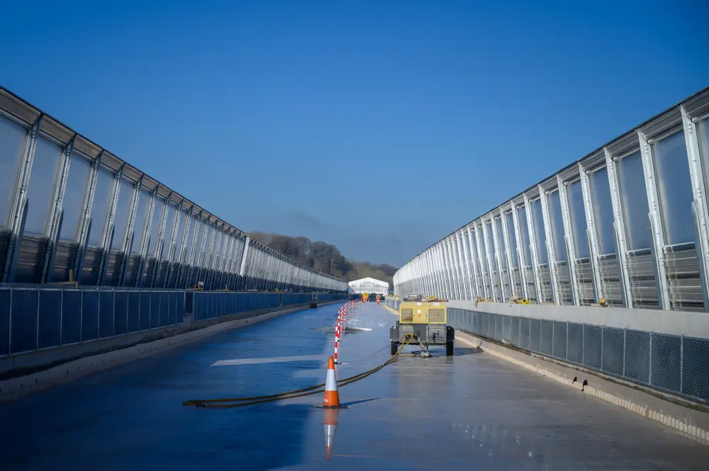 Construction scene on a viaduct with tall, partially transparent noise barriers on both sides. The concrete surface is wet and reflective, with traffic cones and equipment placed along the center. Workers in high-visibility clothing are visible in the distance under a clear blue sky.