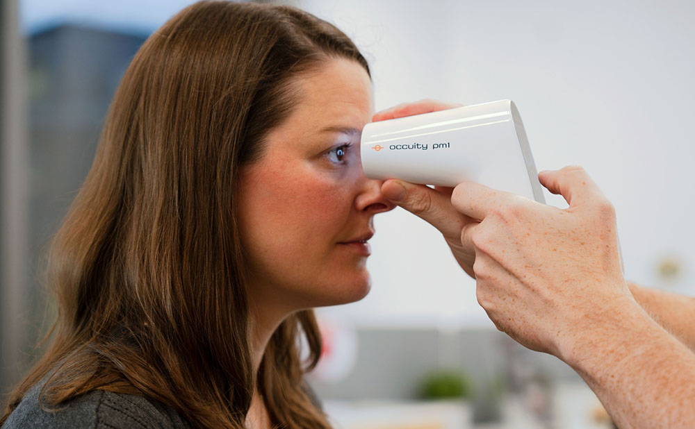 A person is having their eye area scanned with a handheld medical device labelled “Occuity PM1”. The device is white and sleek, held close to the side of the head