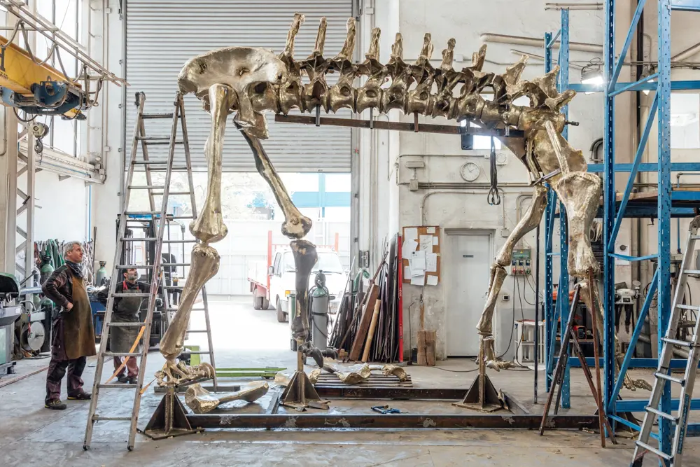 Two men stand in a workshop, regarding a half built bronze sauropod statue