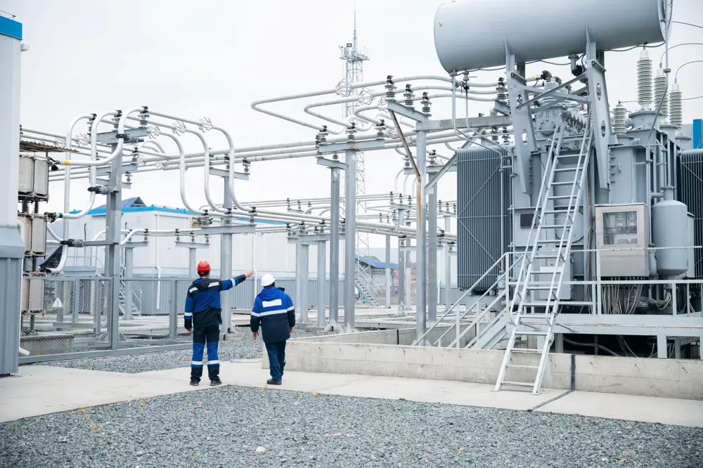 Two engineers in protective clothing walking through an outdoor electrical substation with large transformers and overhead cables, highlighting critical infrastructure.
