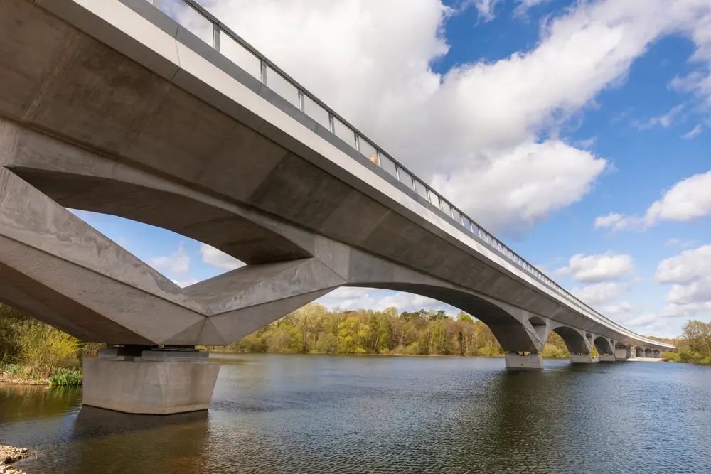 Wide view of a large concrete viaduct spanning a river. The bridge has multiple arches and a sleek design, with noise barriers along the top edge. Trees and greenery line the riverbanks, and the sky is bright with scattered clouds.