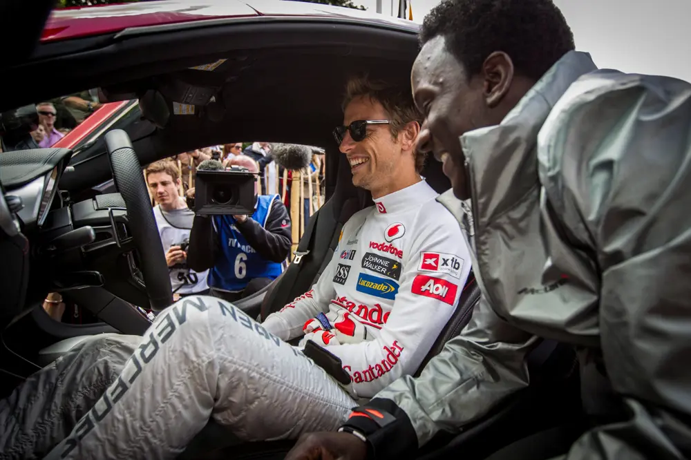 Jenson Button, in a McLaren race suit, sits in a car smiling while talking to James Dornor, who is leaning in through the open door, with photographers nearby.