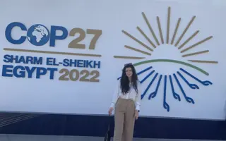 A woman standing in front of a sign for COP27 that says "Sharm El-Sheikh, Egypt 2022"