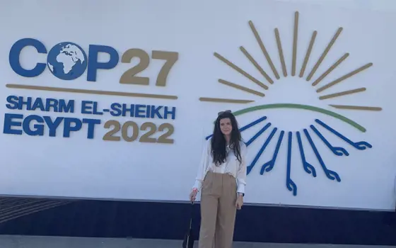A woman standing in front of a sign for COP27 that says "Sharm El-Sheikh, Egypt 2022"