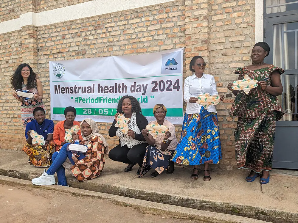 Group of people posing outside a brick building, holding reusable fabric pads in front of a banner reading “Menstrual health day 2024 – #PeriodFriendlyWorld”