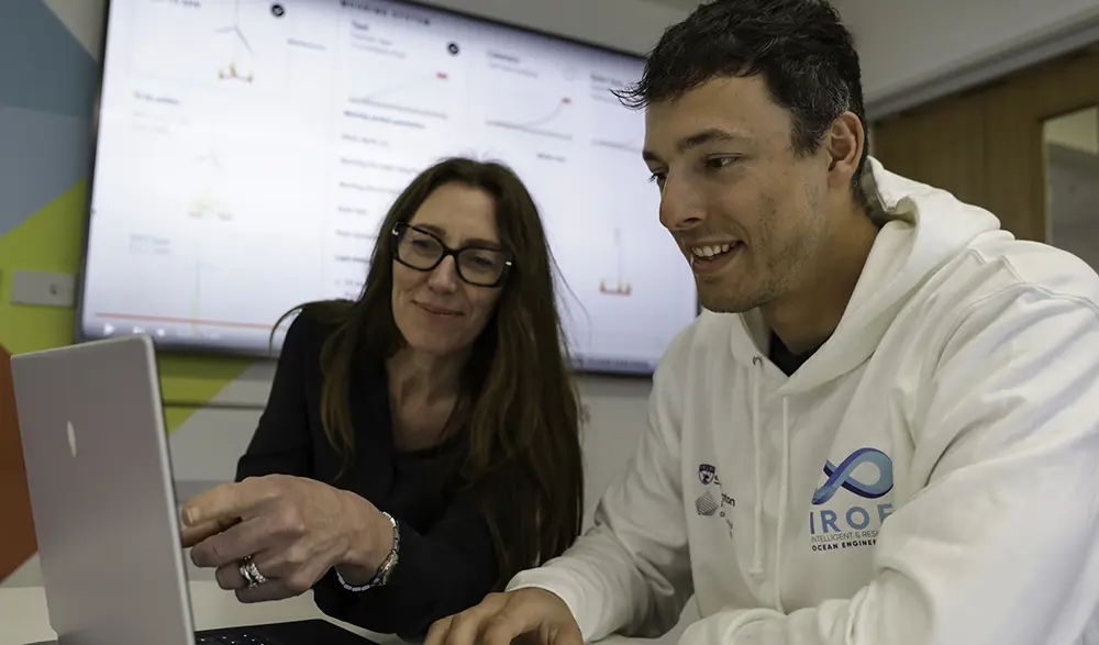 A woman with long dark hair wearing glasses and a dark shirt and a man wearing a white hooded jumper look at a laptop screen