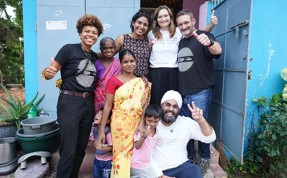 Group of adults and children standing together outside a building, smiling and giving thumbs up next to household equipment and containers