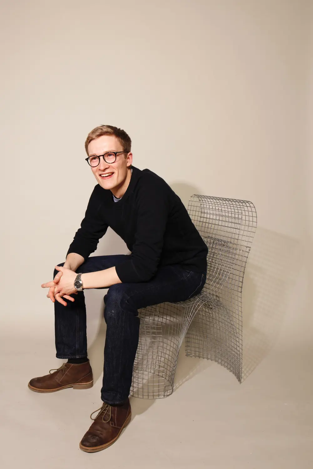 A man with glasses, wearing black, sitting on a chair made of wiring.