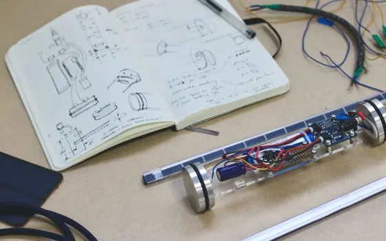 An open book with hand-drawn diagrams and writing, next to a prototype electronic device with colourful cabling exposed.