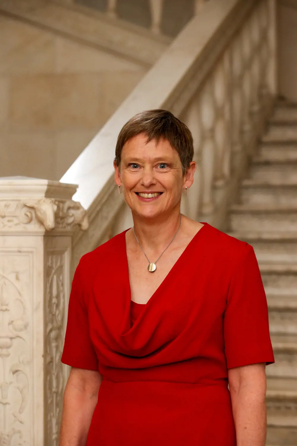 A woman with short hair wearing a red cowl-necked dress, smiling standing on a marble staircase