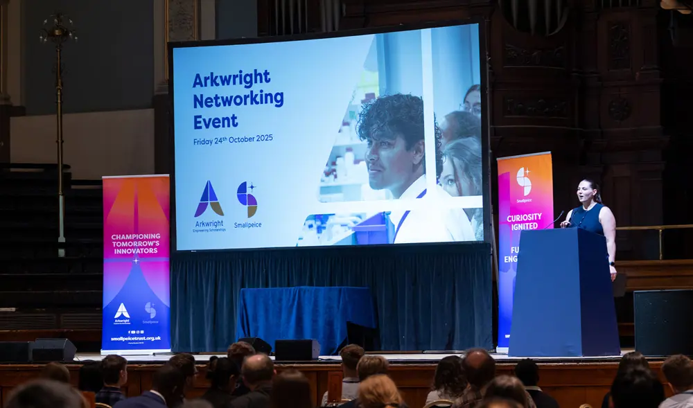 Speaker presenting at a lectern on stage beside a large screen displaying “Arkwright Networking Event” to a seated audience. 