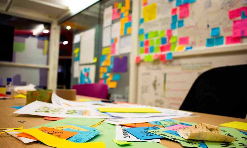 A desk filled with different coloured post-it notes with a whiteboard and more post-it notes in the background. 