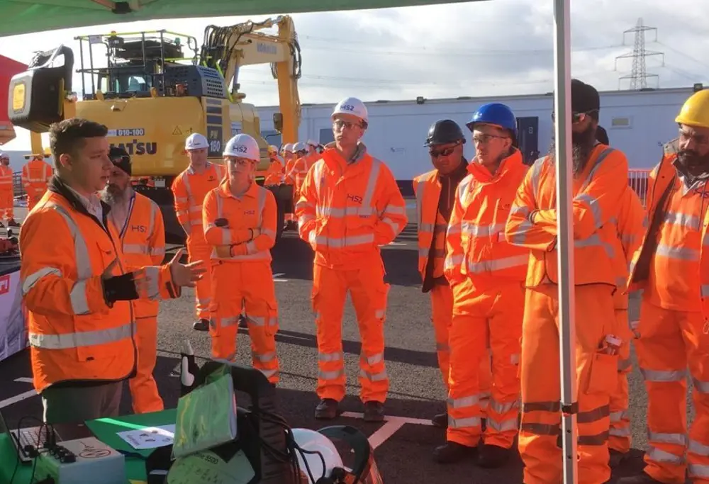 A group of construction workers in high‑visibility orange gear stands in a circle outdoors while another worker gives a demonstration under a canopy, with construction equipment in the background.