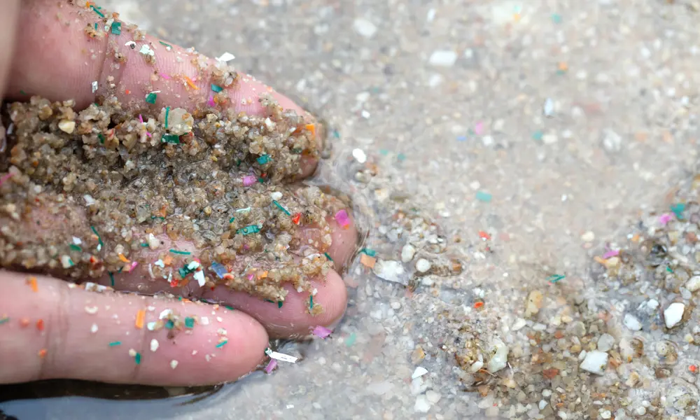 Image shows the fingers from a hand, which is slightly in water having scooped up grains of sand that contain small colourful microplastics