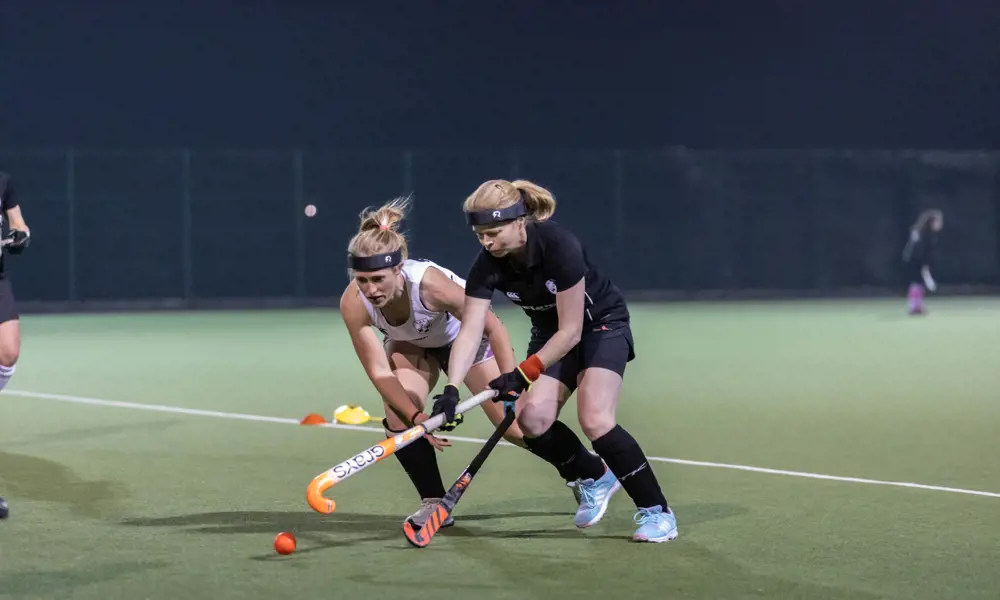 Two white woman hockey players with blonde hair tied in ponytails, aim their hockey sticks towards an orange ball on a green artificial pitch. One is wearing a white sports kit and one is wearing a black sports kit. Both are wearing shinpad and black socks, as well as a protective black sports headband.