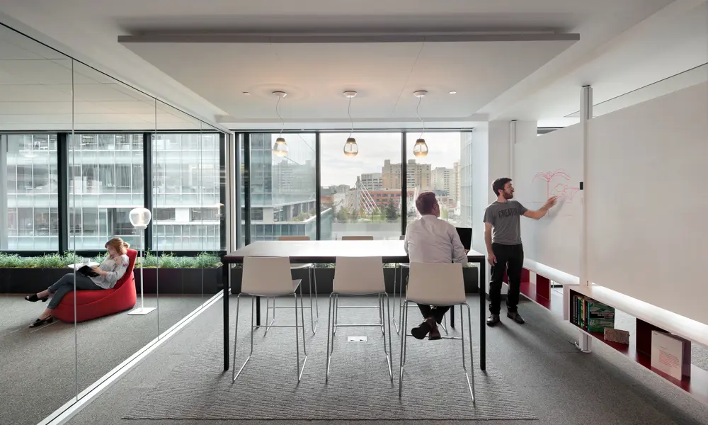 A brightly lit office in San Francisco with one person pointing at a whiteboard and another sitting in a high chair at a table in the same room.