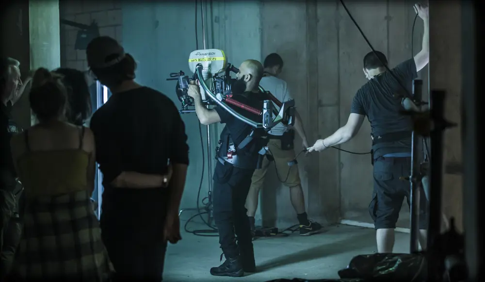 A camera operator wearing a body-mounted stabilizing rig and holding a large professional film camera on a set with concrete walls, cables, and crew members adjusting equipment in the background.