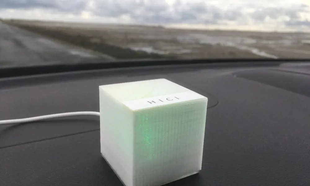 The HICI box that is lit up with green and placed on the dashboard of a car, with a blurry countryside road in the background.