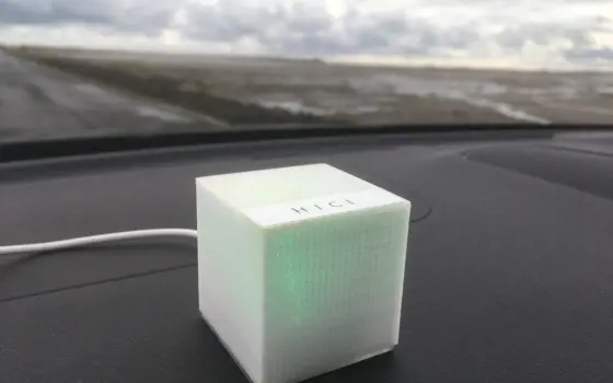 The HICI box that is lit up with green and placed on the dashboard of a car, with a blurry countryside road in the background.