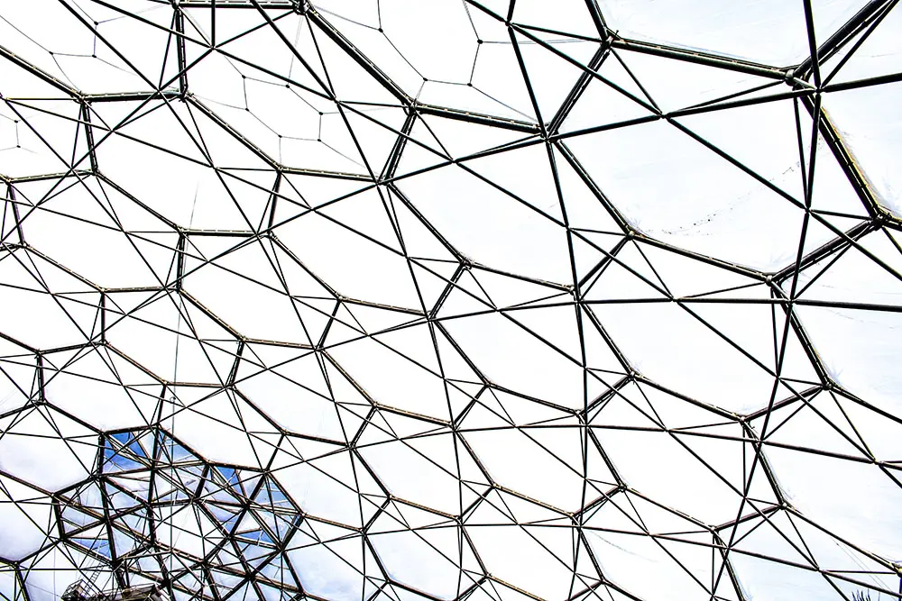 A close‑up view of an intricate geodesic dome structure, showing a network of metal beams forming hexagonal and triangular patterns against the bright sky.