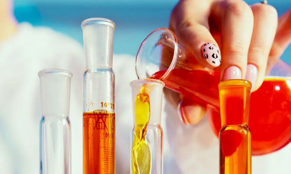 A manicured hand pouring chemicals into lab glassware.