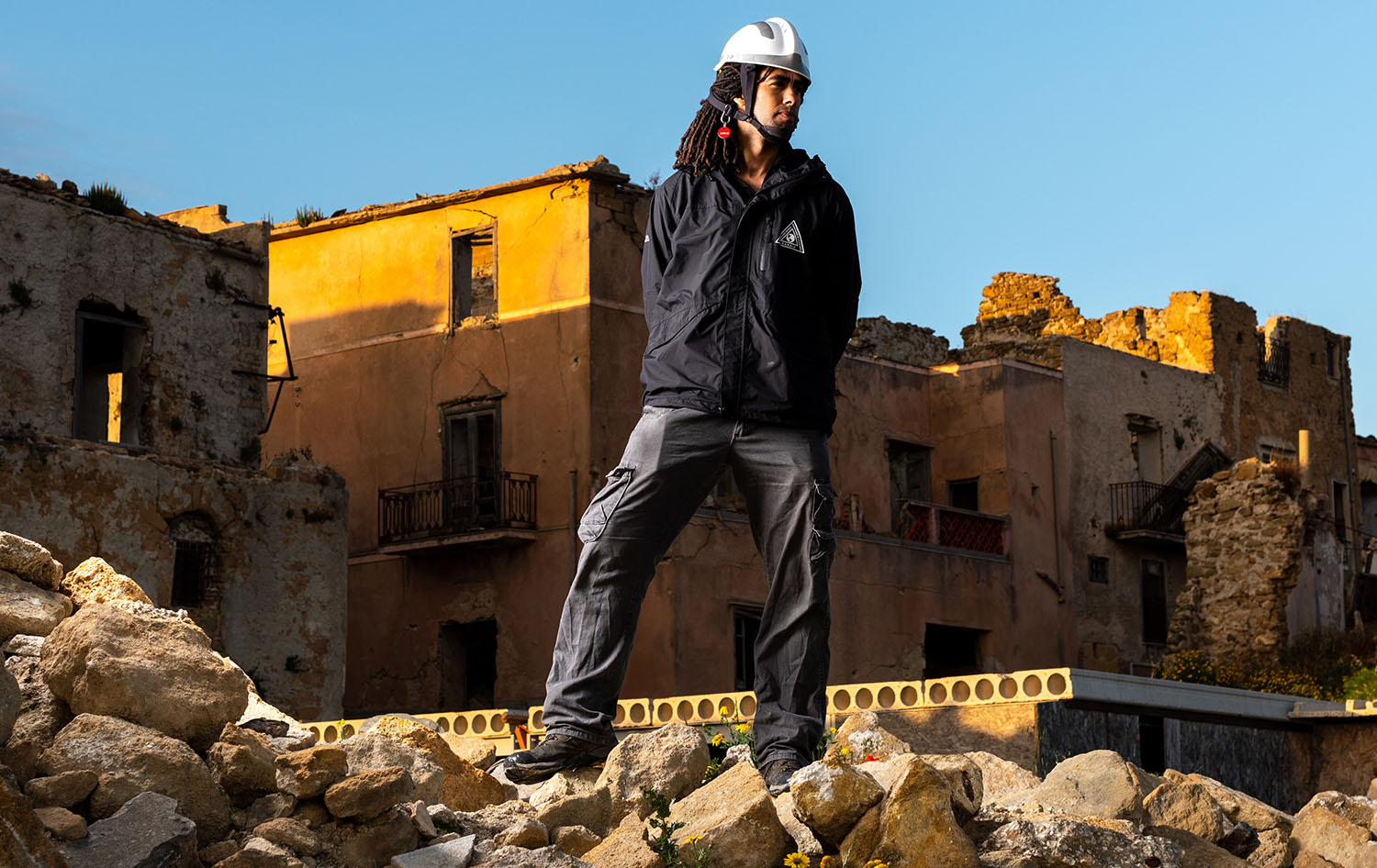 A person wearing outdoor gear and a safety helmet stands on a pile of rubble in front of damaged, sunlit buildings.